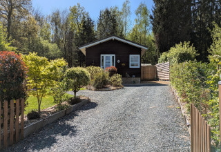 Ingang naar lodge 'Chatillon 67' in België met grindpad, groene planten en houten huisje op de achtergrond.