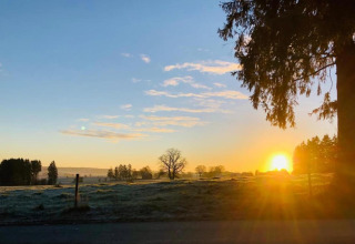 Zonsopgang boven een vredige weide bij een glamping-accommodatie, bomen en een blauwe lucht.