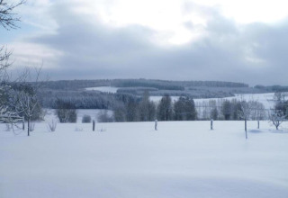 Vue d'hiver d'un hébergement glamping avec champs, arbres et collines enneigés sous un ciel nuageux.