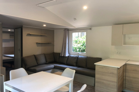 Soggiorno moderno nello chalet Goudhaantje in Belgio con divano angolare grigio, tavolo da pranzo e cucina chiara.