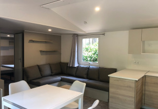 Soggiorno moderno nello chalet Goudhaantje in Belgio con divano angolare grigio, tavolo da pranzo e cucina chiara.