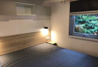 Chambre moderne de chalet avec tête de lit bois, lampe murale et fenêtre sur la nature, en Belgique.