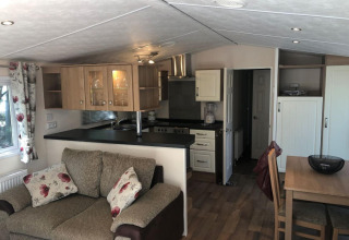 Stue og køkken i Chalet Koperwiek i Belgien, med beige sofa, træbord, gardiner og moderne køkken.