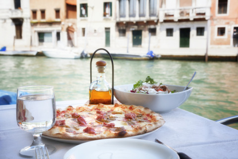 Table with pizza, salad, and water by a canal in Venice, Italy, with historic buildings in the background.
