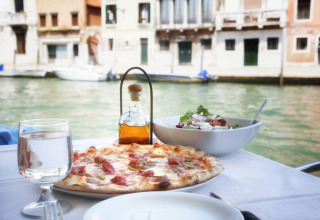 Table with pizza, salad, and water by a canal in Venice, Italy, with historic buildings in the background.