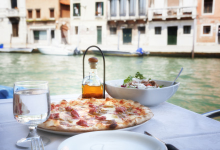 Mesa con pizza, ensalada y agua junto a un canal en Venecia, Italia, con edificios históricos al fondo.