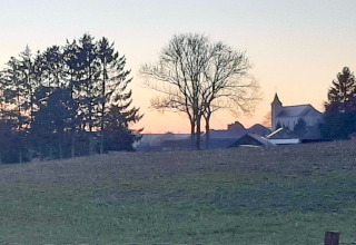 Zonsondergang over een groene weide met bomen en een kerk, gezien vanaf een glampingverblijf.