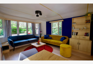 Salon dans le lodge Sijs en Belgique avec canapé bleu, canapé jaune, grande fenêtre et sol en bois.