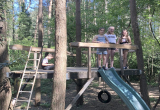 Bambini che giocano su una piattaforma di legno con scivolo in un bosco vicino a un lodge in una giornata di sole.