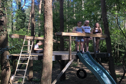 Des enfants jouent sur une plateforme en bois avec toboggan dans une forêt près d’un lodge, en plein soleil.