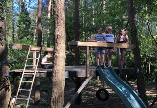 Kinder spielen auf einer Holzplattform mit Rutsche im Wald bei einer Lodge an einem sonnigen Tag.