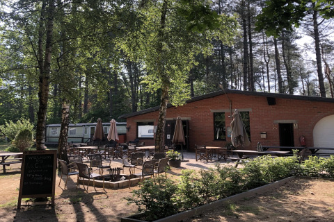 Area glamping all’aperto con tavoli e sedie davanti a una struttura in mattoni, circondata da alberi.