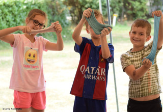 Tre bambini sorridenti giocano con lo slime colorato all’aperto in un lodge, mostrando le loro creazioni.