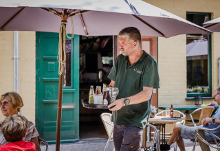 A waiter serves drinks to guests sitting outdoors under umbrellas at a glamping accommodation.