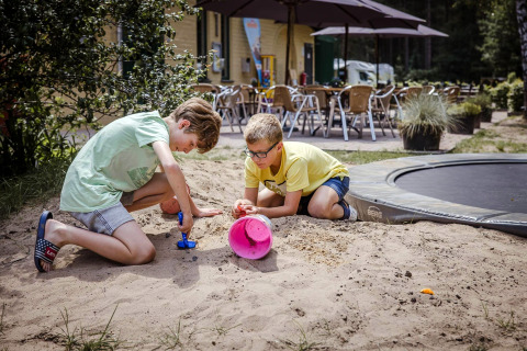 Deux garçons jouent avec des jouets dans le sable près d'un lodge avec café en terrasse et parasols.