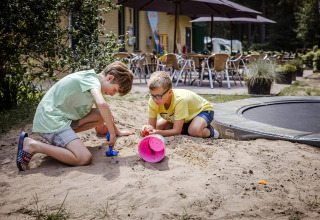 Dos niños juegan en la arena con juguetes cerca de un alojamiento con cafetería al aire libre y sombrillas.
