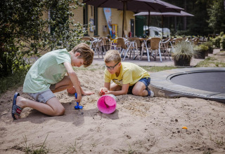 Deux garçons jouent avec des jouets dans le sable près d'un lodge avec café en terrasse et parasols.