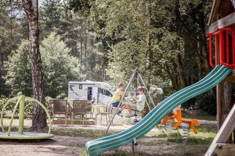 Kinderen spelen op een speeltuin bij een glampingplek met een mobilhome op de achtergrond in het groen.