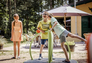 Trois enfants jouent dehors près d’un lodge, deux garçons rient sur un manège pendant qu’une fille observe.