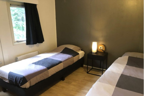 Chambre confortable au lodge 'Ekster' en Belgique avec deux lits simples, table de nuit et fenêtre avec rideau.