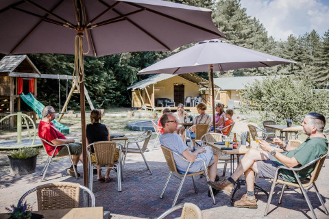 Persone sedute sotto ombrelloni in un caffè all'aperto di un lodge con parco giochi e bosco sullo sfondo.