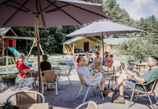 Personer sidder under parasoller på en udendørs café ved en lodge med legeplads og skov i baggrunden.