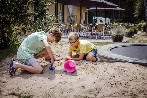 Deux enfants jouent dans un bac à sable avec des jouets, près d’une terrasse et d’un trampoline à glamping.