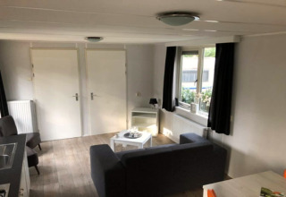 Foto del interior de una acogedora sala en la cabaña Koolmees en Bélgica, con sofá y ventanas.