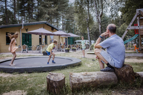 Un homme regarde des enfants jouer sur un trampoline devant un lodge en forêt avec terrasse extérieure.