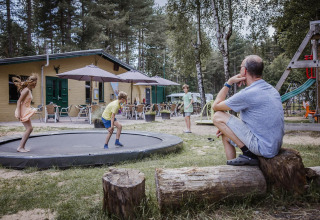 Un homme regarde des enfants jouer sur un trampoline devant un lodge en forêt avec terrasse extérieure.