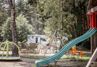 Twee kinderen spelen op een schommel op een speeltuin bij een camper en terrasstoelen in het groen.