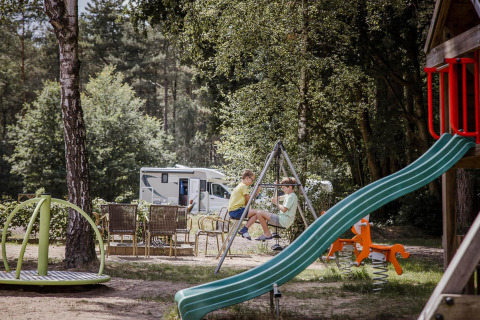 Two children play on swings at a playground near a camper, surrounded by trees and outdoor seating.