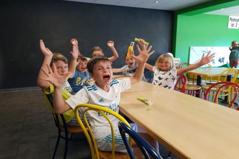 Kinderen zitten aan een lange tafel in een lodge, lachen en steken hun handen enthousiast in de lucht.
