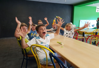 Kinderen zitten aan een lange tafel in een lodge, lachen en steken hun handen enthousiast in de lucht.