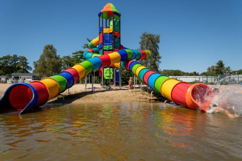 Kleurrijke waterglijbaan bij een lodge, kinderen glijden een meer in op een zonnige heldere dag.