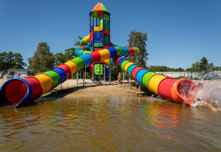 Kleurrijke waterglijbaan bij een lodge, kinderen glijden een meer in op een zonnige heldere dag.