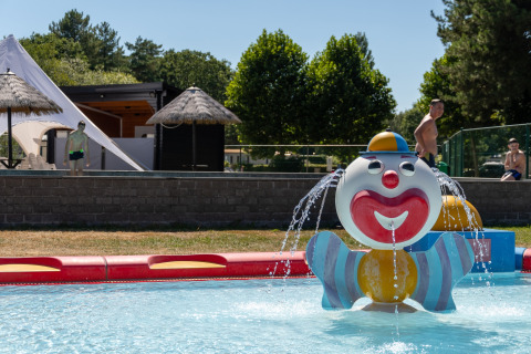 Kleurrijke clownfontein in kinderbad bij een lodge, met kinderen, volwassenen en tenten op de achtergrond.