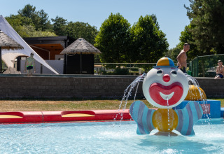 Kleurrijke clownfontein in kinderbad bij een lodge, met kinderen, volwassenen en tenten op de achtergrond.
