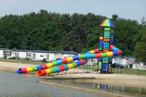 Kleurrijke glijbaan aan het strand bij een lodge, met chalets en bomen op de achtergrond, zonnige dag.