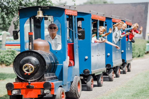 Kinderen en een oudere man maken een ritje in een blauwe minitrein bij een lodge en zwaaien vrolijk.