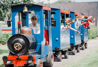 Bambini e un uomo anziano si divertono su un trenino blu in un lodge, sorridendo e salutando dal finestrino.