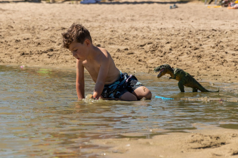 Jongen speelt aan de waterrand bij het strand van een lodge, met een speelgoed dinosaurus achter hem.