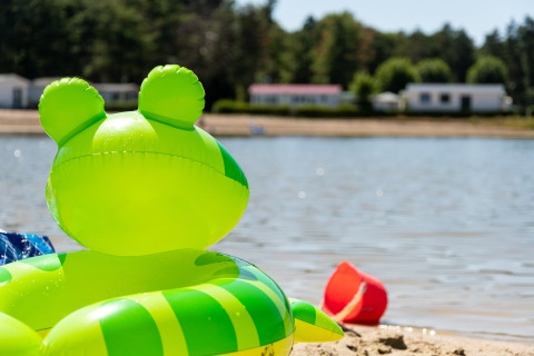Groene opblaasband en rode emmer op zandstrand bij een meer met huisjes op de achtergrond.