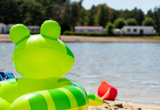 Groene opblaasband en rode emmer op zandstrand bij een meer met huisjes op de achtergrond.