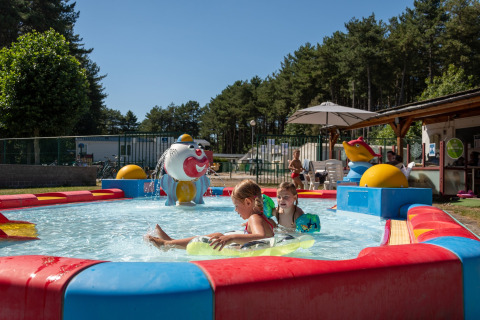 Bambini giocano in una piscina bassa colorata vicino a un lodge circondato da alberi e natura.