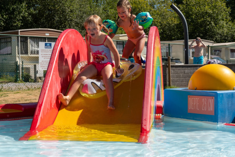 Due bambini giocano su uno scivolo d'acqua colorato in un lodge con case mobili e sole sullo sfondo.