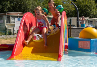 Twee kinderen spelen op een kleurrijke waterglijbaan bij een lodge met stacaravans op een zonnige dag.
