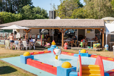 Mensen ontspannen aan tafels op het overdekte terras naast een kinderbadje bij een lodge op een zonnige dag.