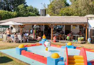 Mensen ontspannen aan tafels op het overdekte terras naast een kinderbadje bij een lodge op een zonnige dag.