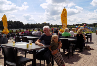 Een man en een kind zitten samen op het terras van een lodge met een speeltuin op de achtergrond.